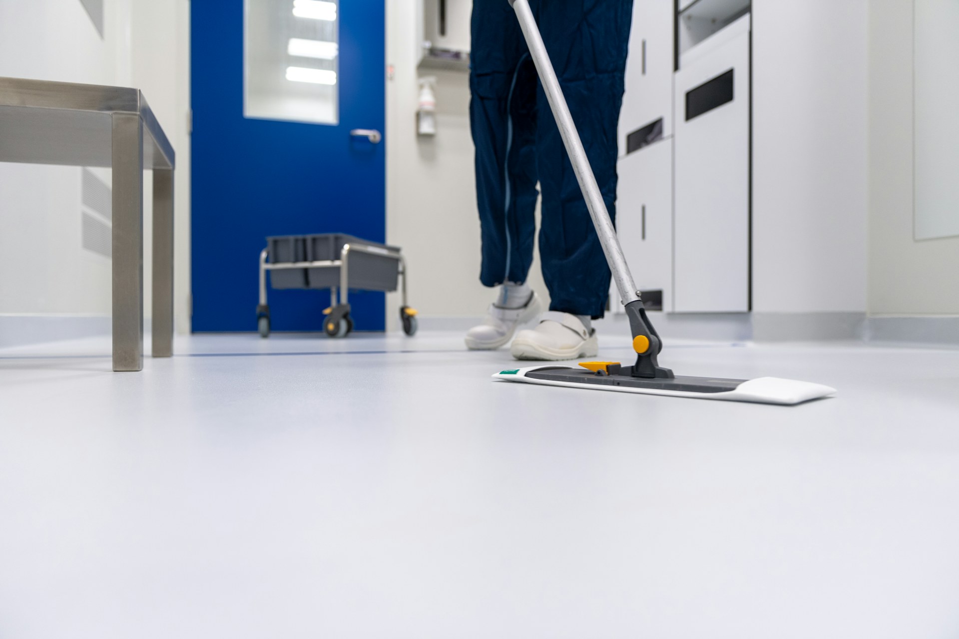 A cleaner in blue workwear mops a white floor in a long, bright corridor