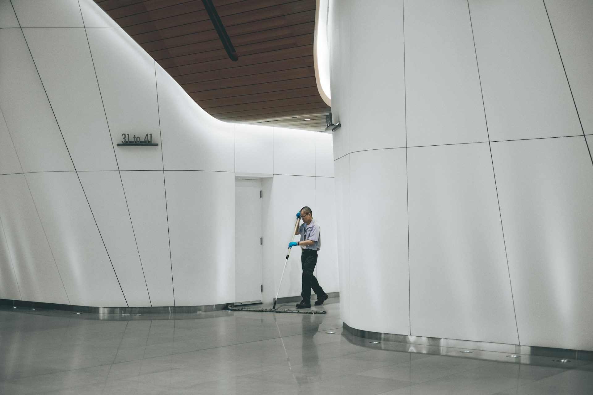A man cleans the floor beside a plain white wall in a minimal, well-lit interior