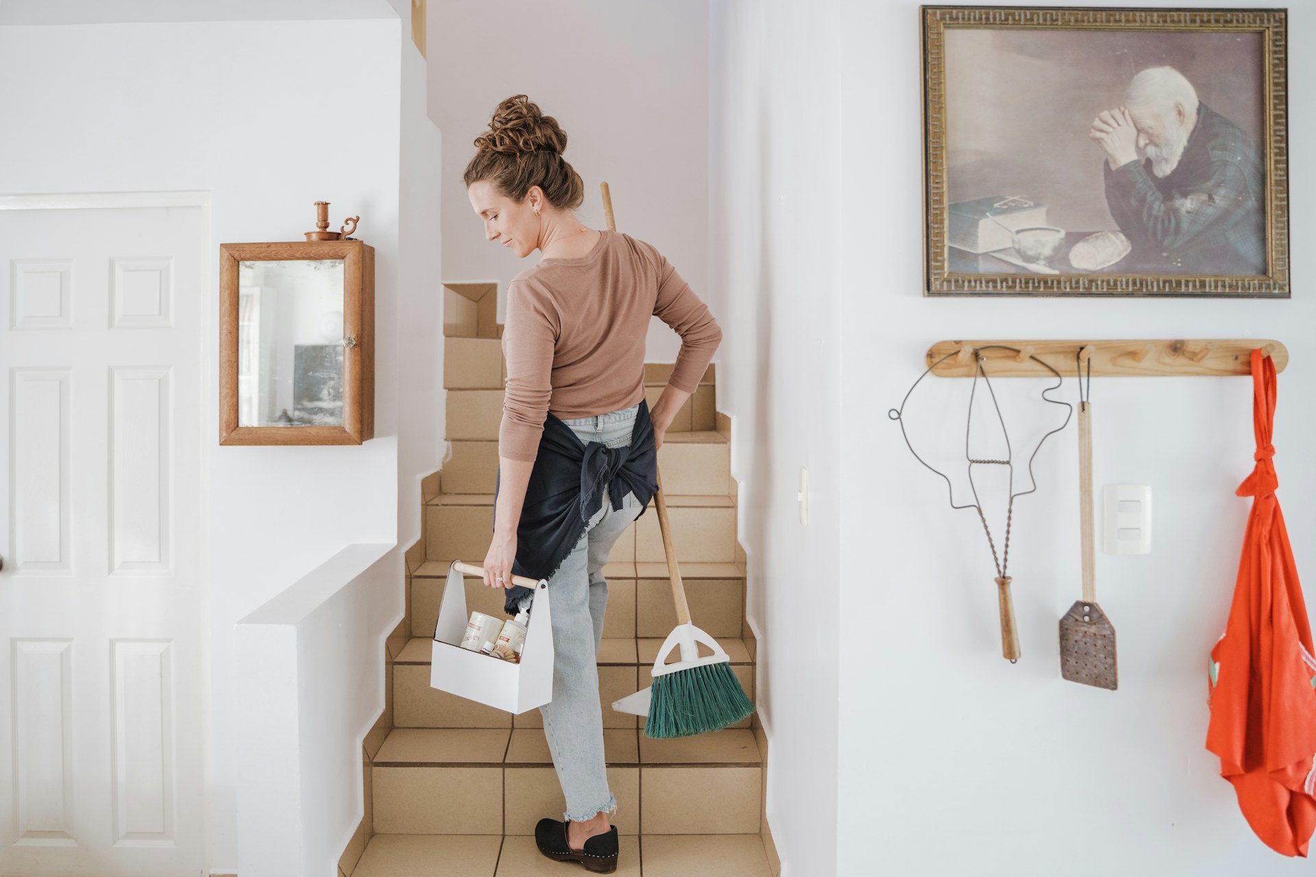 A cleaner carries a cleaning caddy and broom up tiled stairs in a home